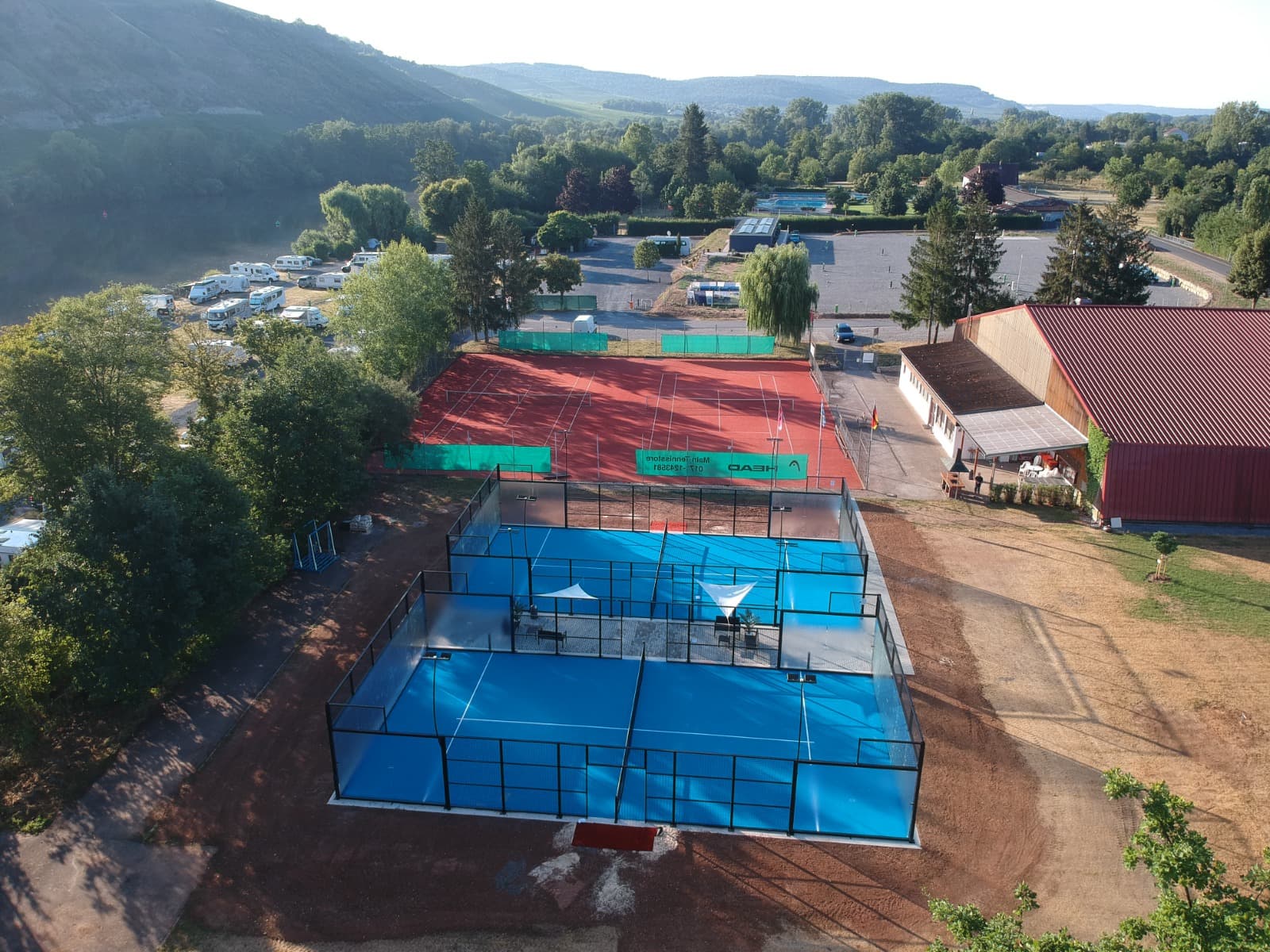 Blick von oben auf die beiden blauen Padelcourts, den roten Tennisplatz und die Vereinshalle des TV Zellingen